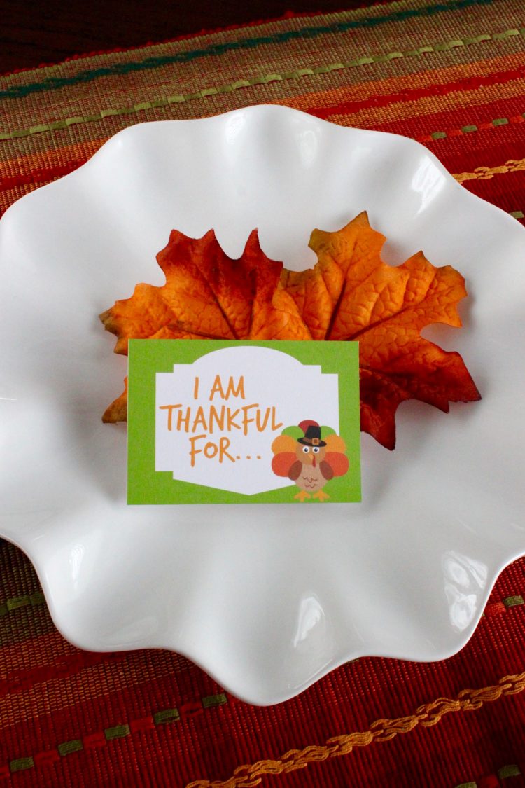 Dress up the Thanksgiving table with these free printable Thanksgiving Conversation Starters! I am thankful for card is placed on a few silk fall leaves on a white plate creating a festive table setting.