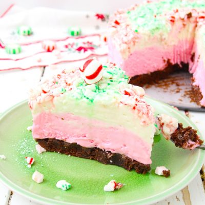 Beautiful no bake peppermint cheesecake slice on a green plate. Whole cheesecake is featured in the back with peppermint pieces sprinkled around the plate.