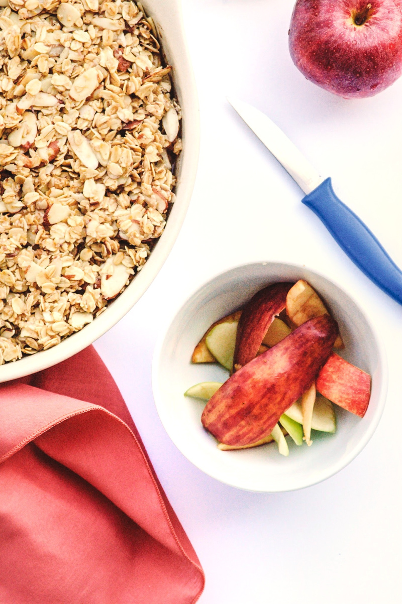 This apple crisp is served in a white dish adorn with a amber napkin, cut up apples and a paring knife.