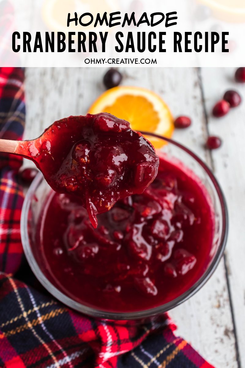 A freshly made bowl of homemade cranberry sauce with a close up of a heaping spoonful. Displayed beautifully on a red plaid table covering and slices of oranges. 