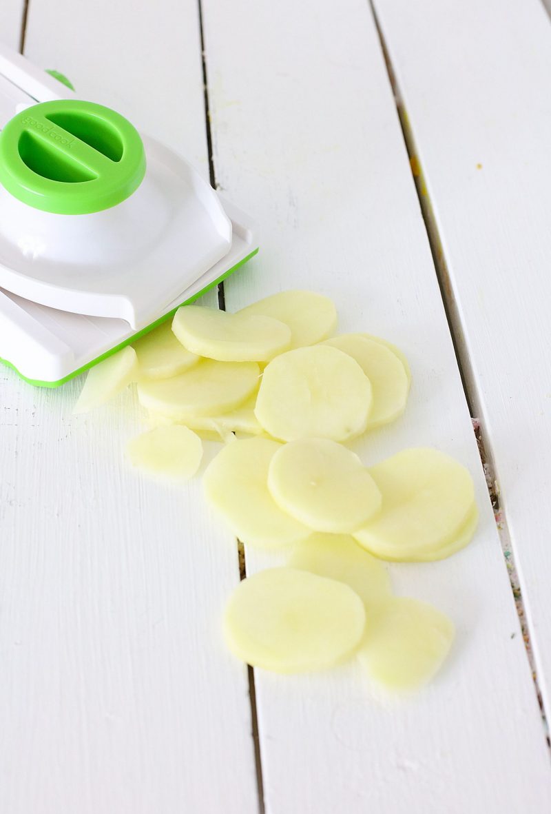 Sliced potatoes and mandolin slicer sitting on a white background. 