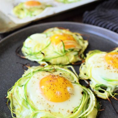 Three zucchini egg nest on a black plate with a baking pan of additional zucchini nests.