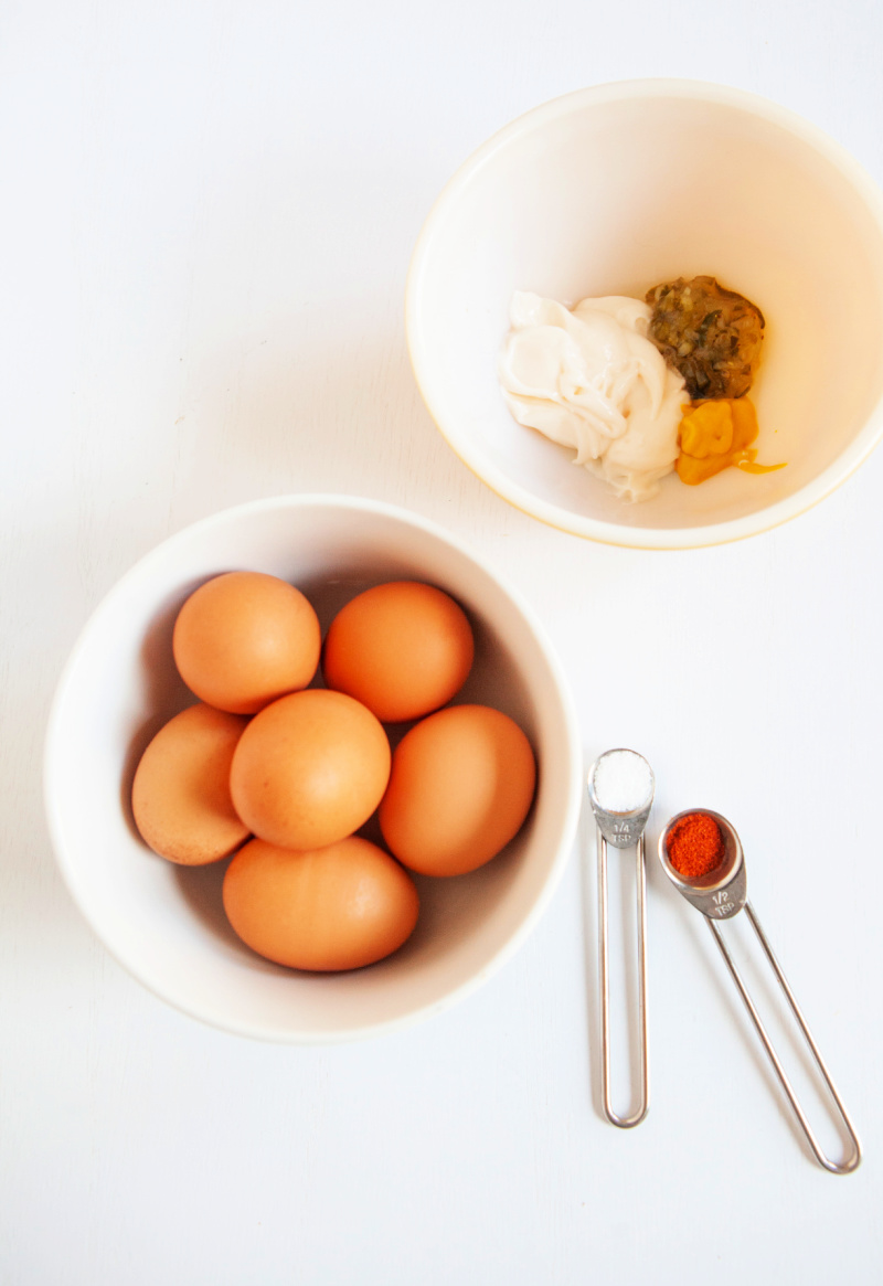 Healthy deviled egg ingredients including a bowl of hard boiled brown eggs, measuring spoons with spices and a white bowl of additional ingredients.