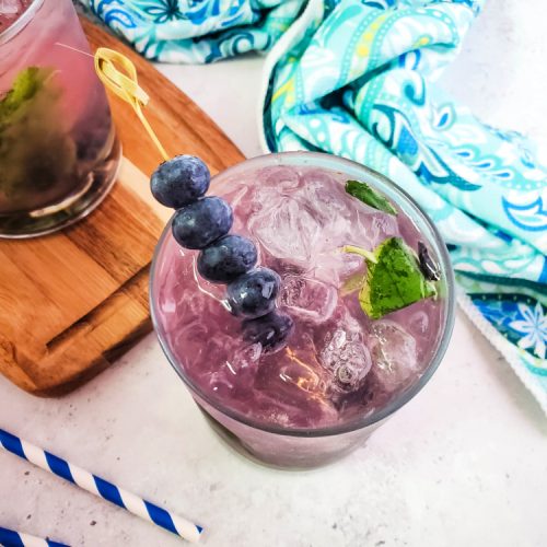 A blueberry mojito cocktail garnished with a skewer of blueberries and surrounded by a aqua hand towel and blue striped straws.