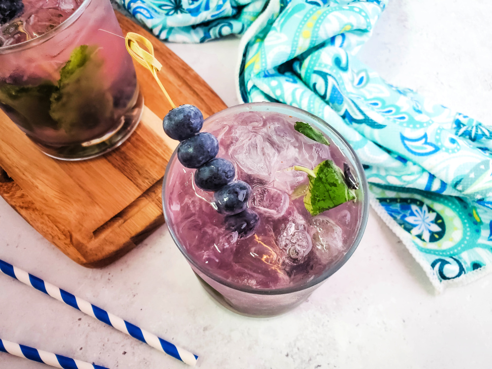 A blueberry mojito cocktail garnished with a skewer of blueberries and surrounded by a aqua hand towel and blue striped straws.