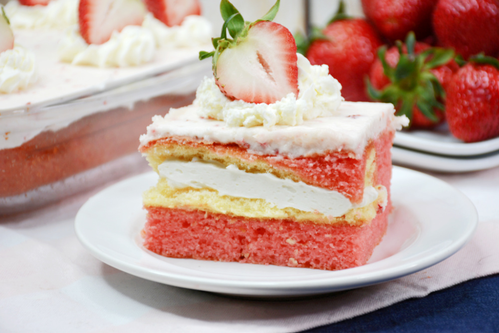 A square slice of strawberry Twinkie cake with homemade frosting and a slice of strawberry on top. The strawberry cake is sitting on a white dessert plate with a dish of strawberries in the background.
