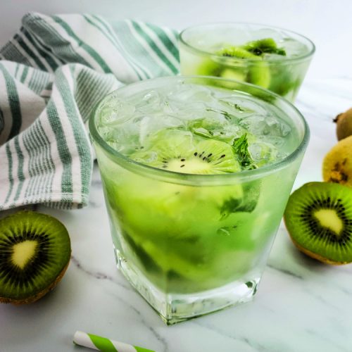 Kiwi mojito drink in a short cocktail glass with sliced kiwi, green and white cloth hand towel and a white marble background.