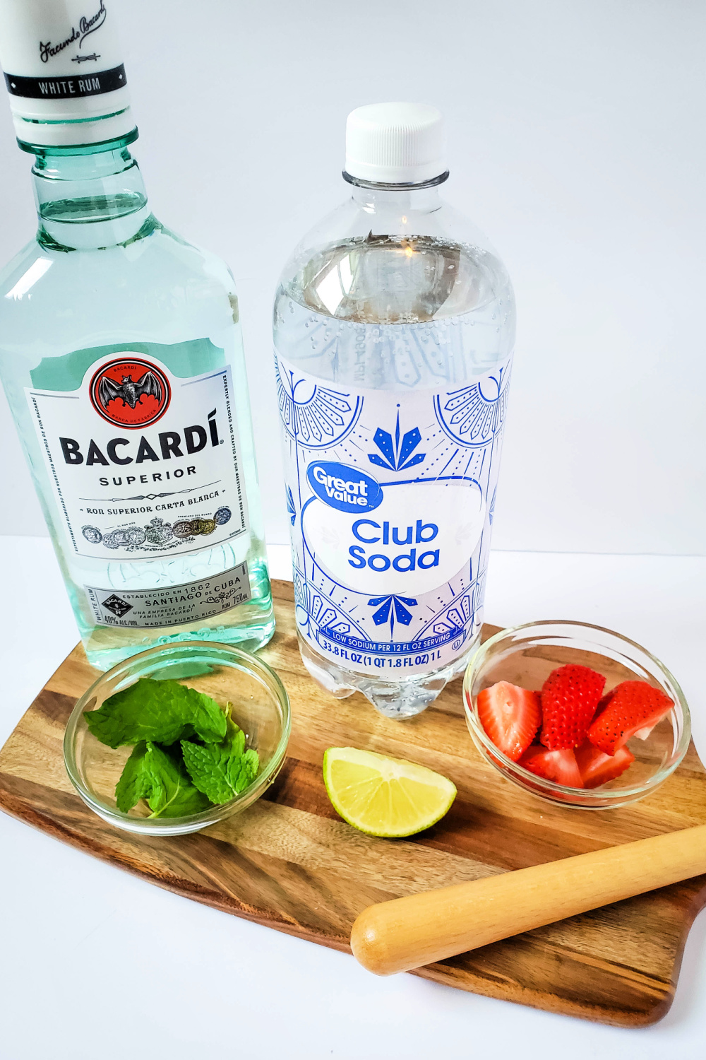 All the ingrediants to make a strawberry mojito. Rum, club soda, mint, strawberries and a slice of lime sitting on a cutting board.