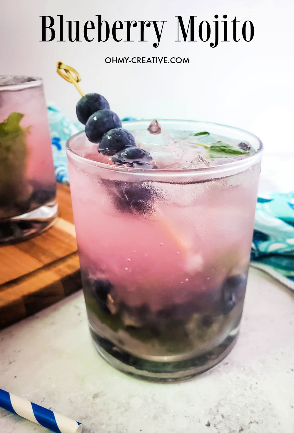 A blueberry mojito cocktail garnished with a skewer of blueberries and surrounded by a aqua hand towel and blue striped straw