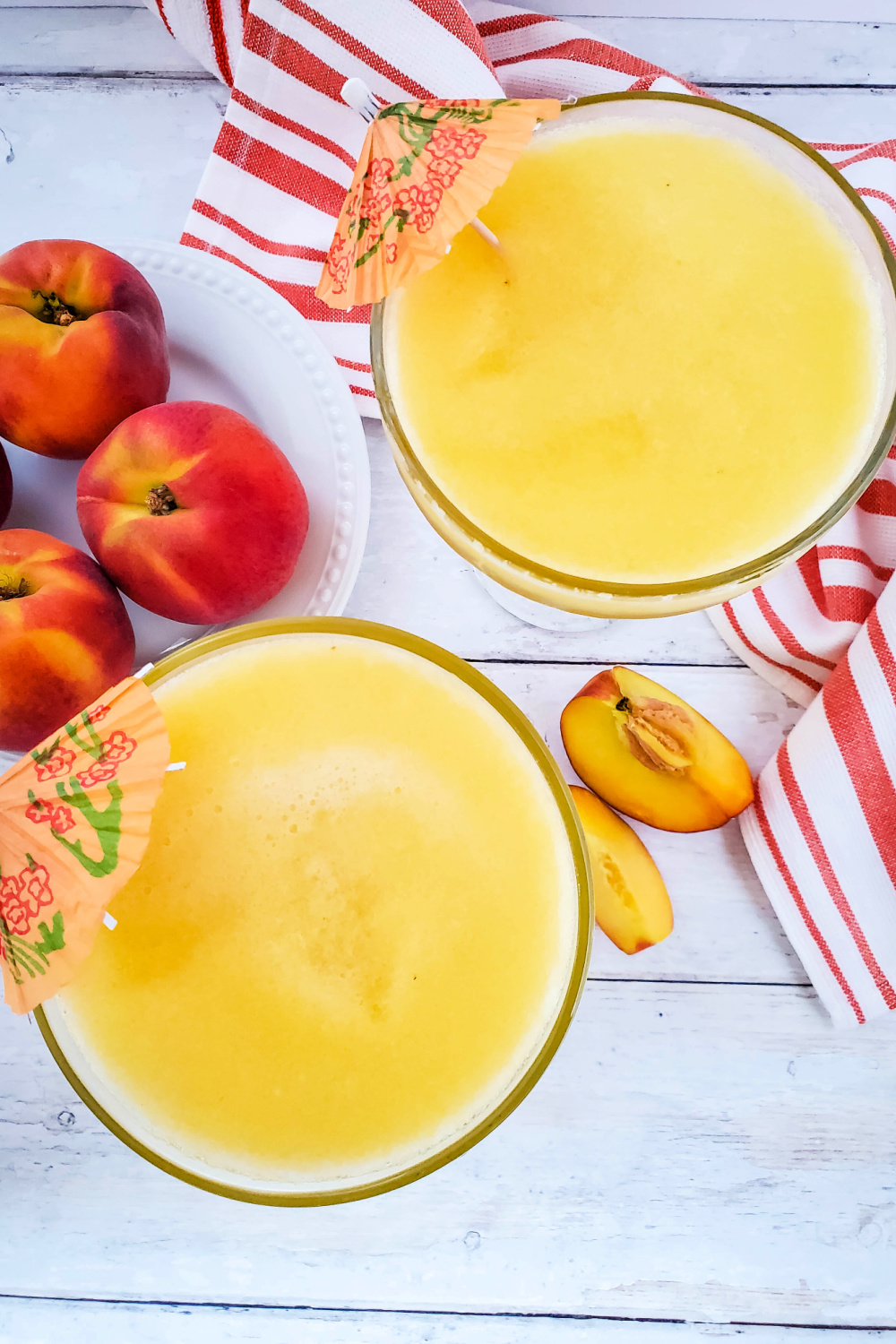 These two frozen peach margaritas are garnished with fun drink umbrellas. They are sitting on a white wood background with a stripped hand towel and sliced peaches. 