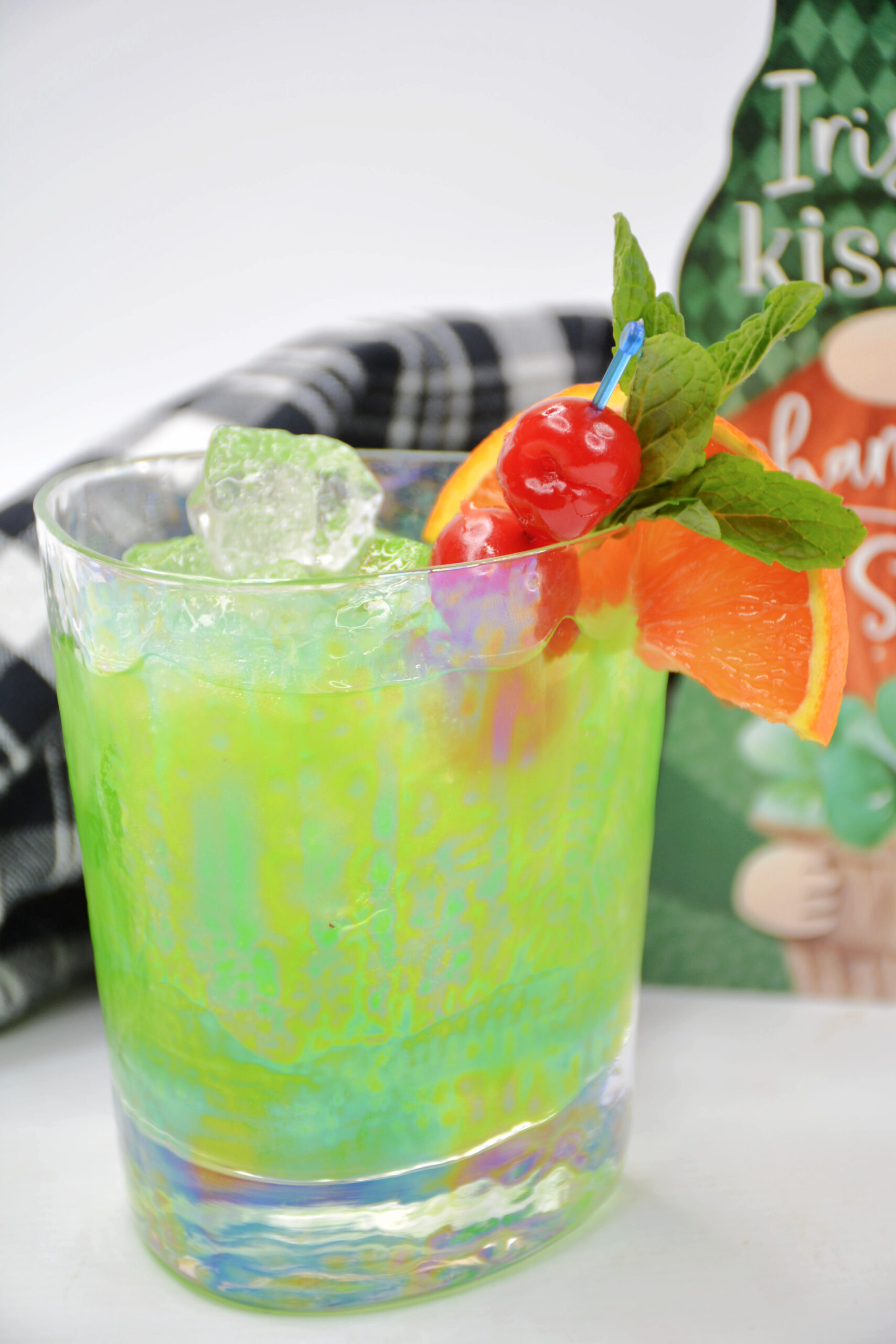 Green cocktail garnished with a slice of orange, mint, and cherries and a leprechaun sign in the background and a black and white napkin.