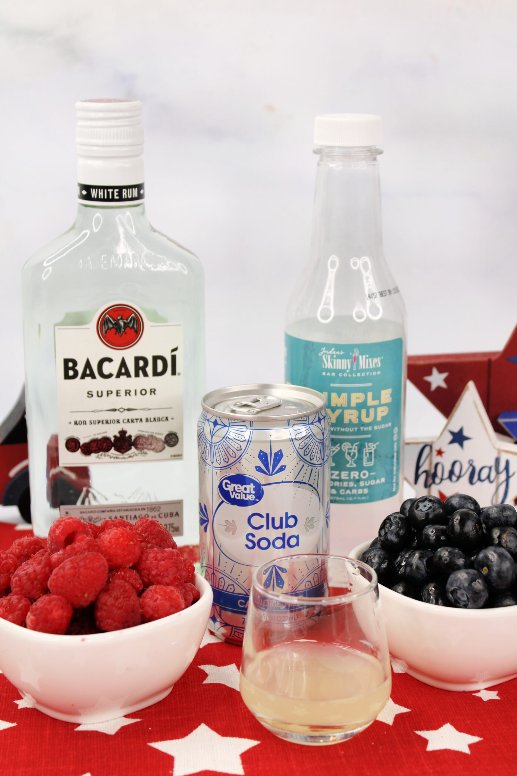 Ingredients for a Fourth of July Mojito include raspberries, blueberries, rum, simple syrup and club soda. These ingredients are sitting on a pretty red cloth napkin with white stars.
