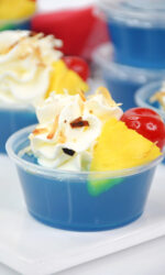 These blue Hawaiian jello shots are made with tasty berry blue jello topped with whipped cream, toasted coconut garnished with pineapple and a maraschino cherry. These jello shots are sitting on a red checked napkin on a white background.