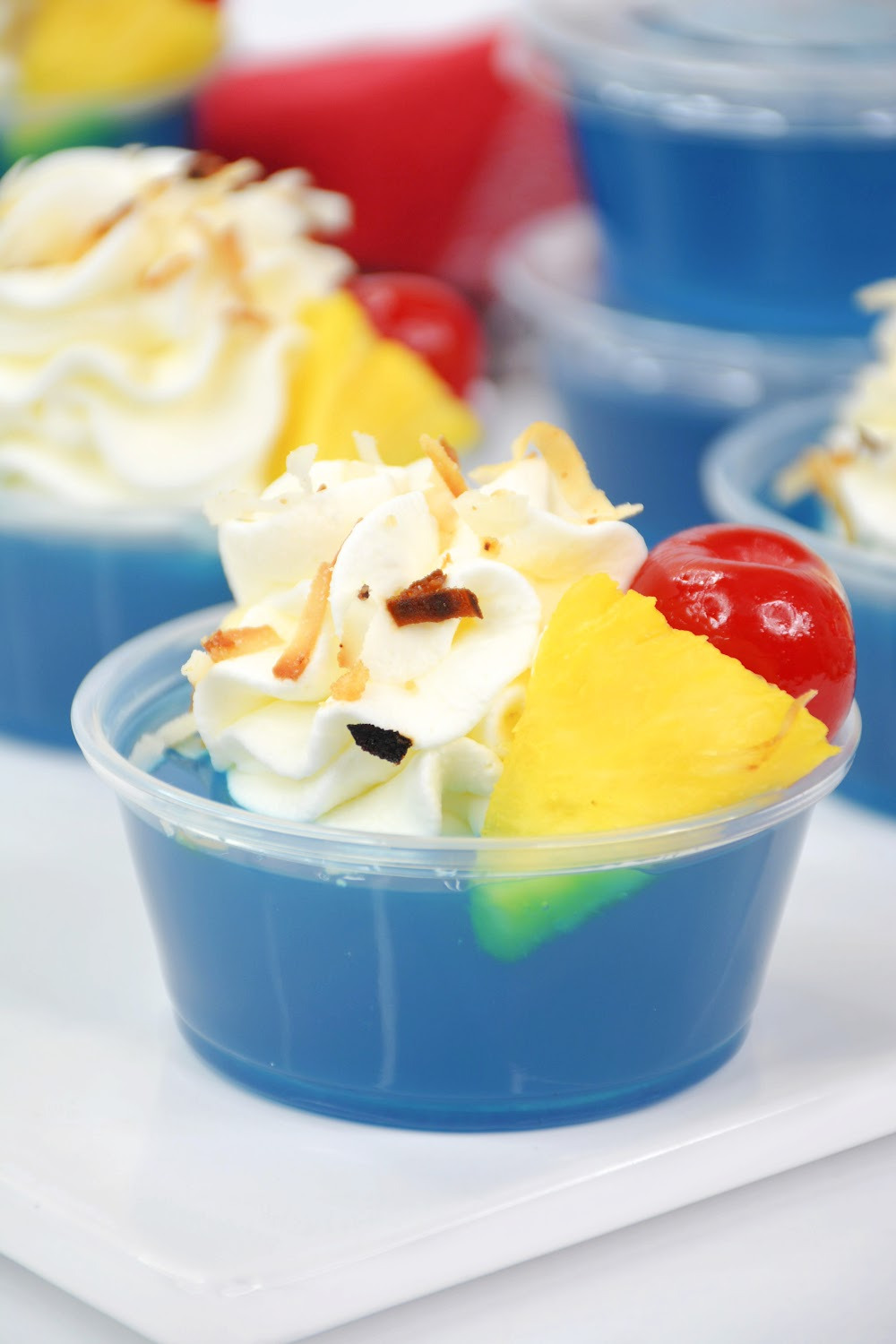 These blue Hawaiian jello shots are made with tasty berry blue jello topped with whipped cream, toasted coconut garnished with pineapple and a maraschino cherry. These jello shots are sitting on a red checked napkin on a white background.