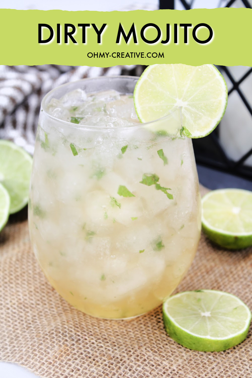 This dirty mojito cocktail is sitting on a burlap background with a black and white checked napkin and a black summer lantern. Garnished include slices of lime.