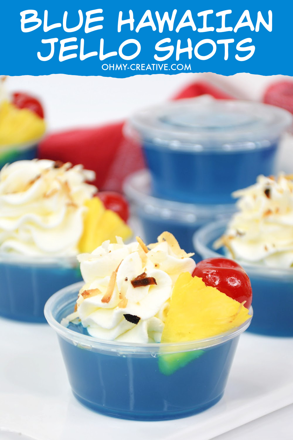 These blue Hawaiian jello shots are made with tasty berry blue jello topped with whipped cream, toasted coconut garnished with pineapple and a maraschino cherry. These jello shots are sitting on a red checked napkin on a white background. 