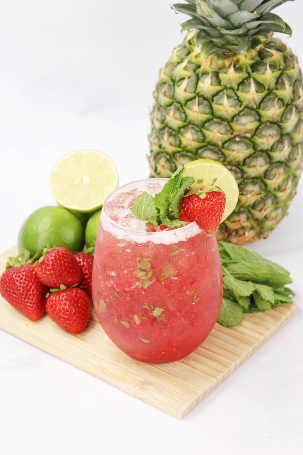 This Strawberry Pineapple Mojito cocktail is garnished with strawberry and mint. It is sitting on a wooden cutting board surrounded by strawberries, limes and a whole pineapple.