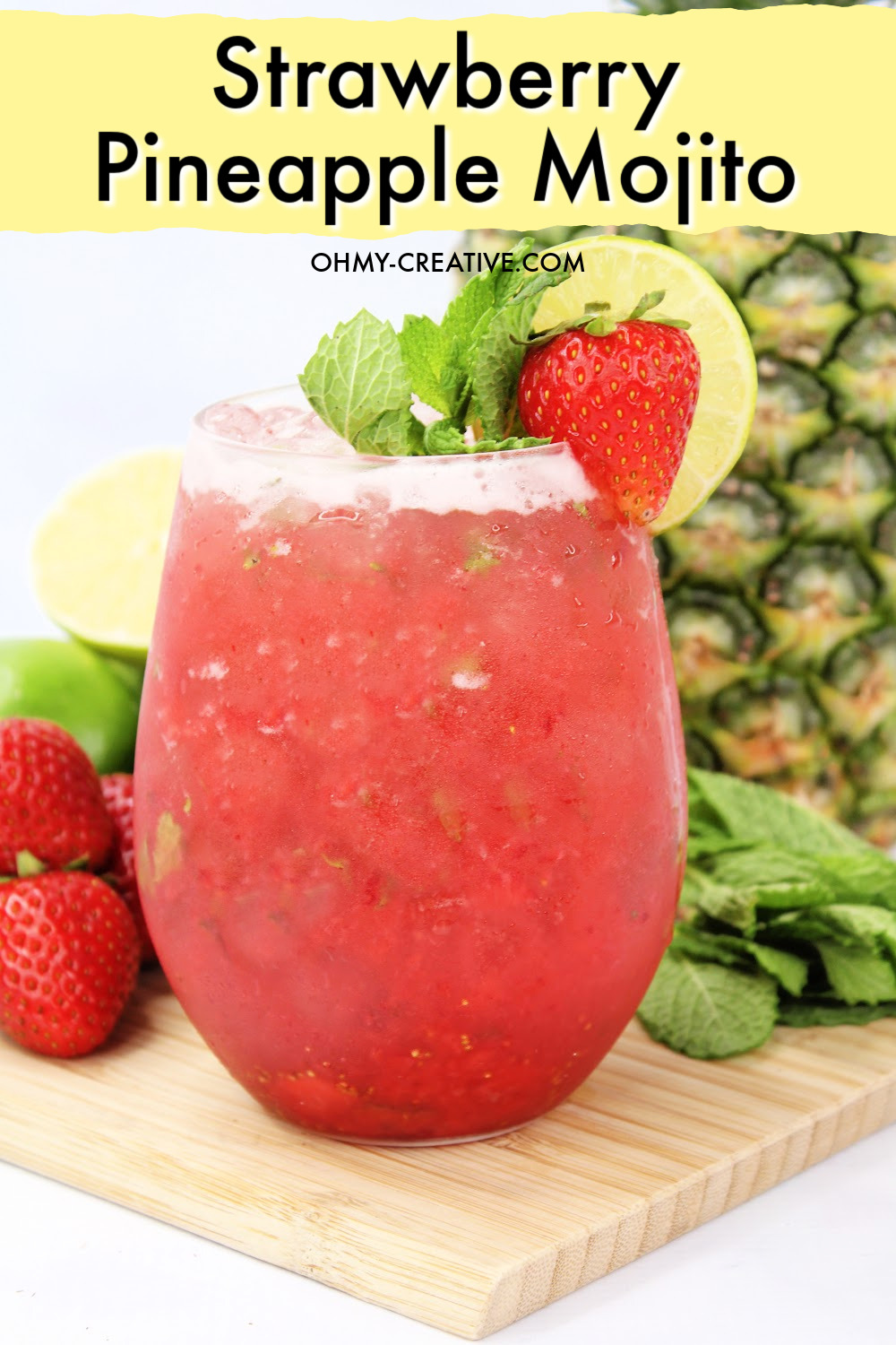 This Strawberry Pineapple Mojito cocktail is garnished with strawberry and mint. It is sitting on a wooden cutting board surrounded by strawberries, limes and a whole pineapple. 