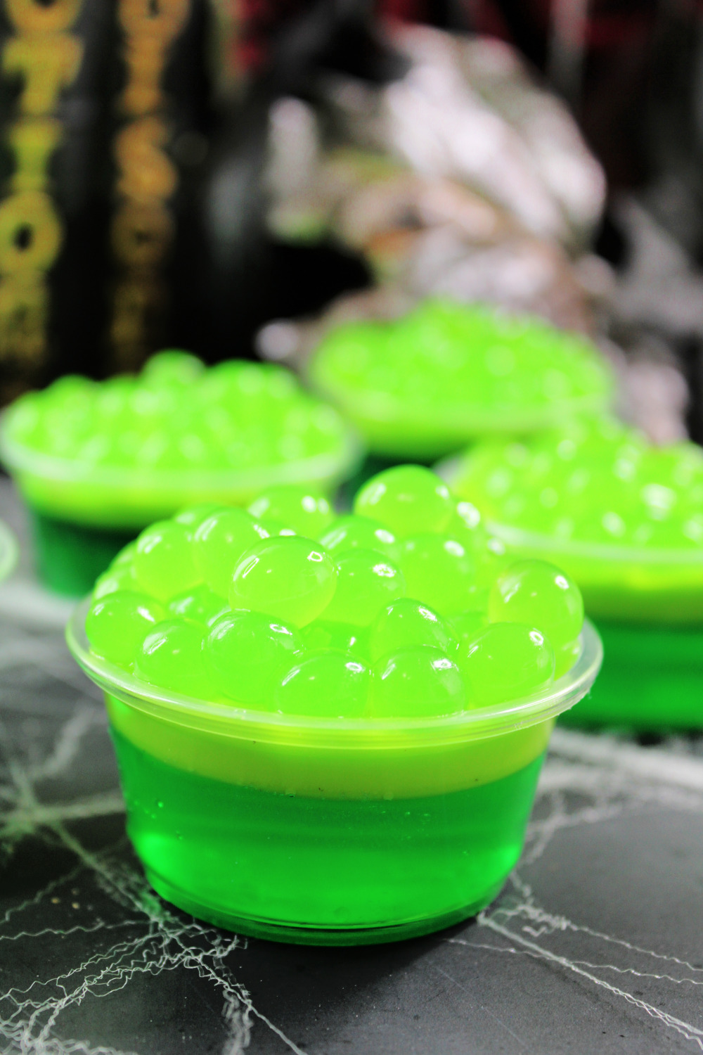 Bubbly Witches Brew Jello Shots for Halloween Parties. These green jello shots are sitting on a spiderweb background. The tops of the jello shots are topped with green apple popping bobas.