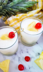 Two glasses of piña colada cocktails sitting on a marble table with pineapples in the background. These piña coladas at garnished with pineapple and maraschino cherries.