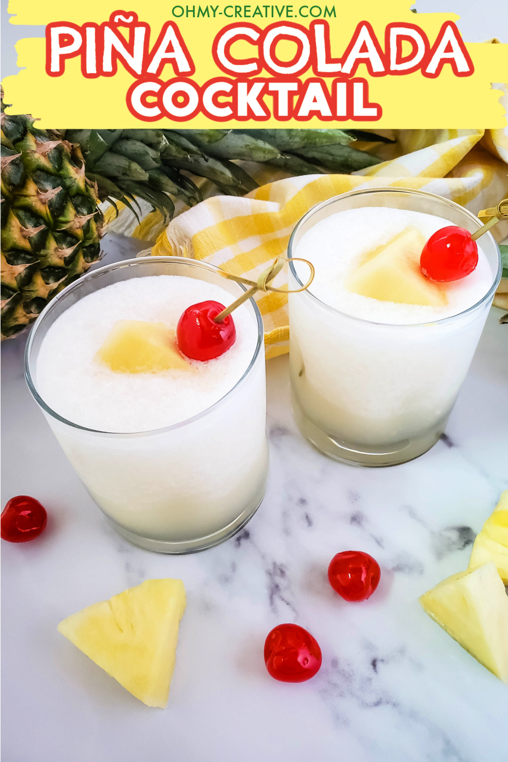 Two glasses of piña colada cocktails sitting on a marble table with pineapples in the background. These piña coladas at garnished with pineapple and maraschino cherries.