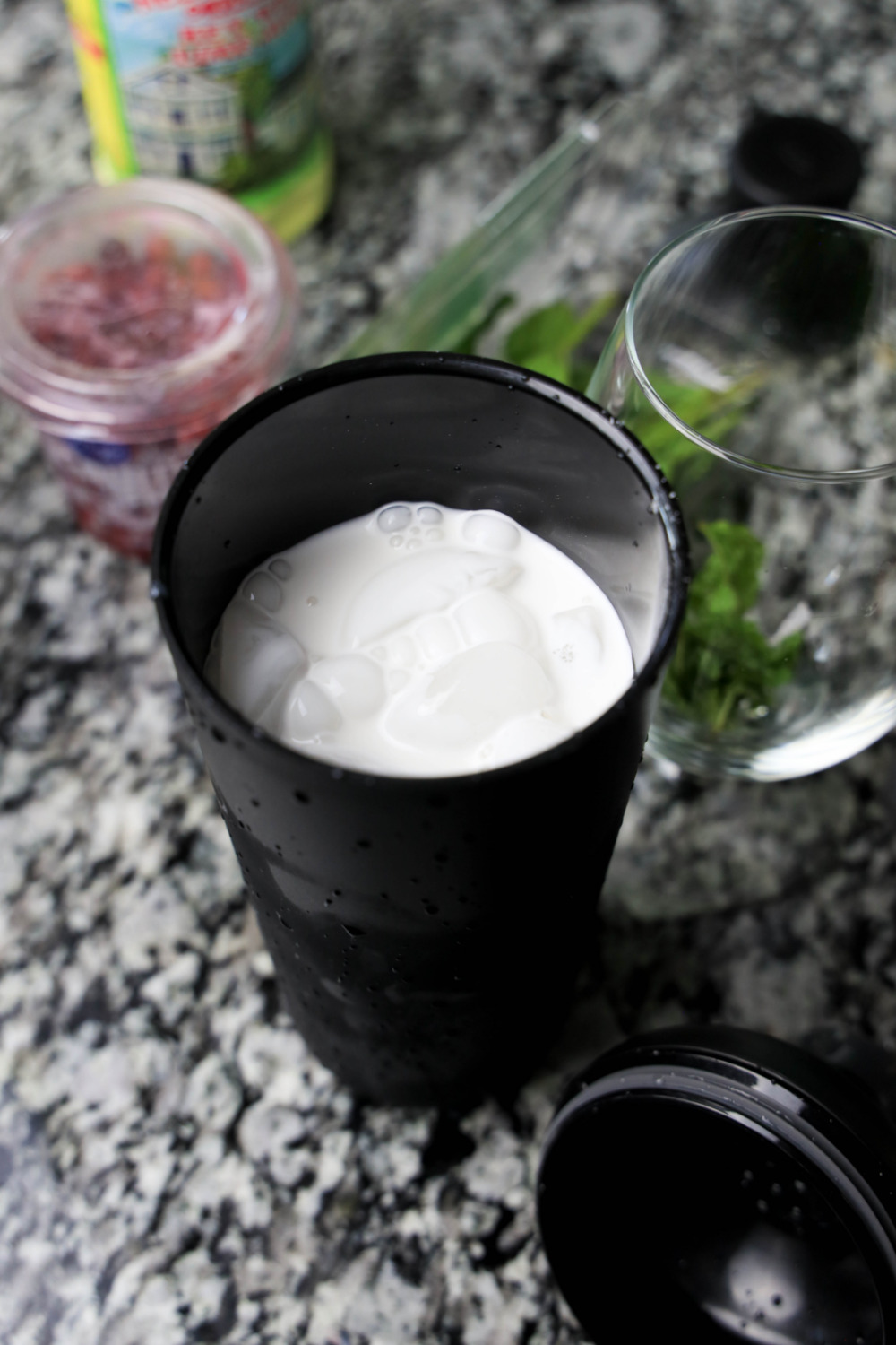 A white Christmas mojito cocktail in a cocktail shaker. It sits on a black and white granite countertop with some of the mojito ingredients around it.