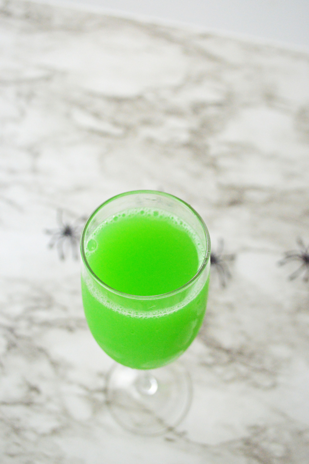 A single glass of orange juice, blue curacao and champagne sitting on the gray and white marble background with spiders. The orange juice looks green with all the ingredients mixed together for the Halloween Mimosa.