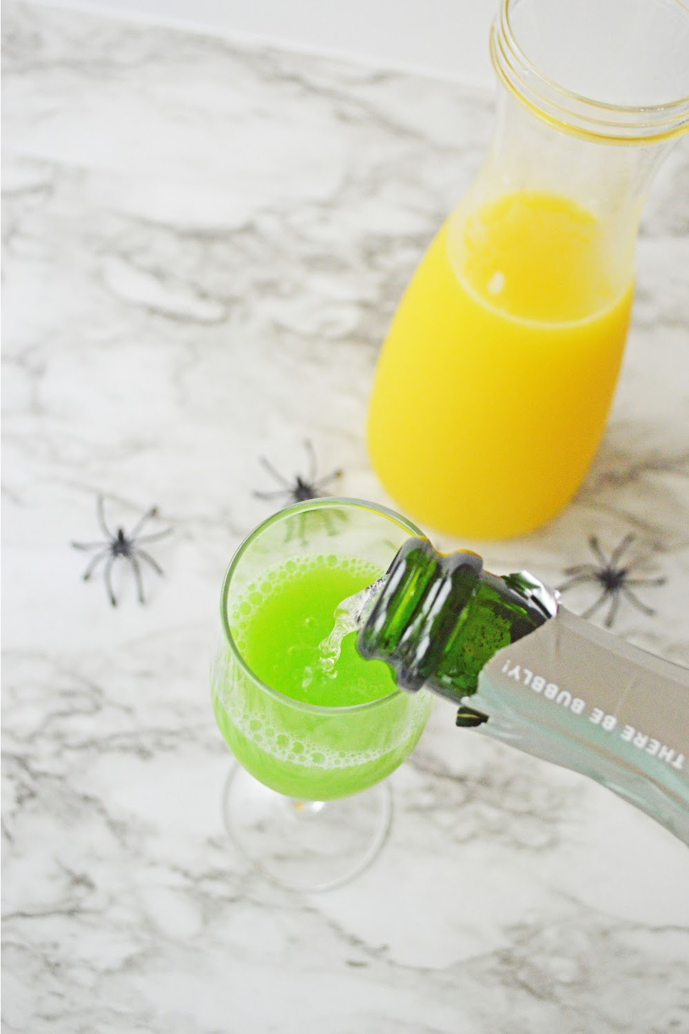 Pouring champagne into the glass that already has orange juice and blue Curacao in it for the Halloween Mimosas ingredients include champagne, orange juice and blue Curacao. These ingredients are displayed on a gray and white marble background with black plastic spiders.
