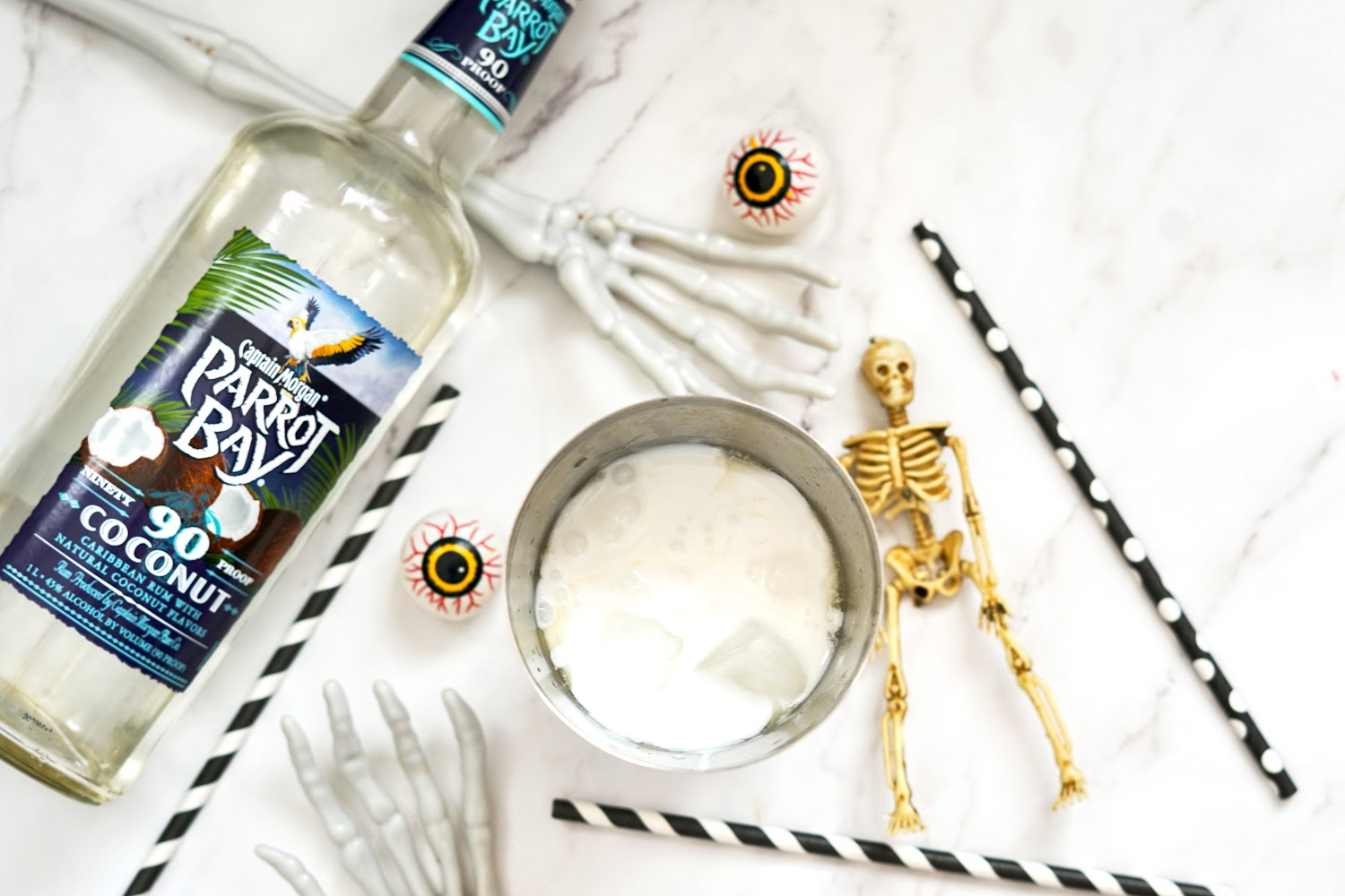 Laying flat on a white marble back ground are the Halloween pina colada mix in a shaker and the bottle of rum. Eyeballs and mini skeletons make this feel a little spooky.