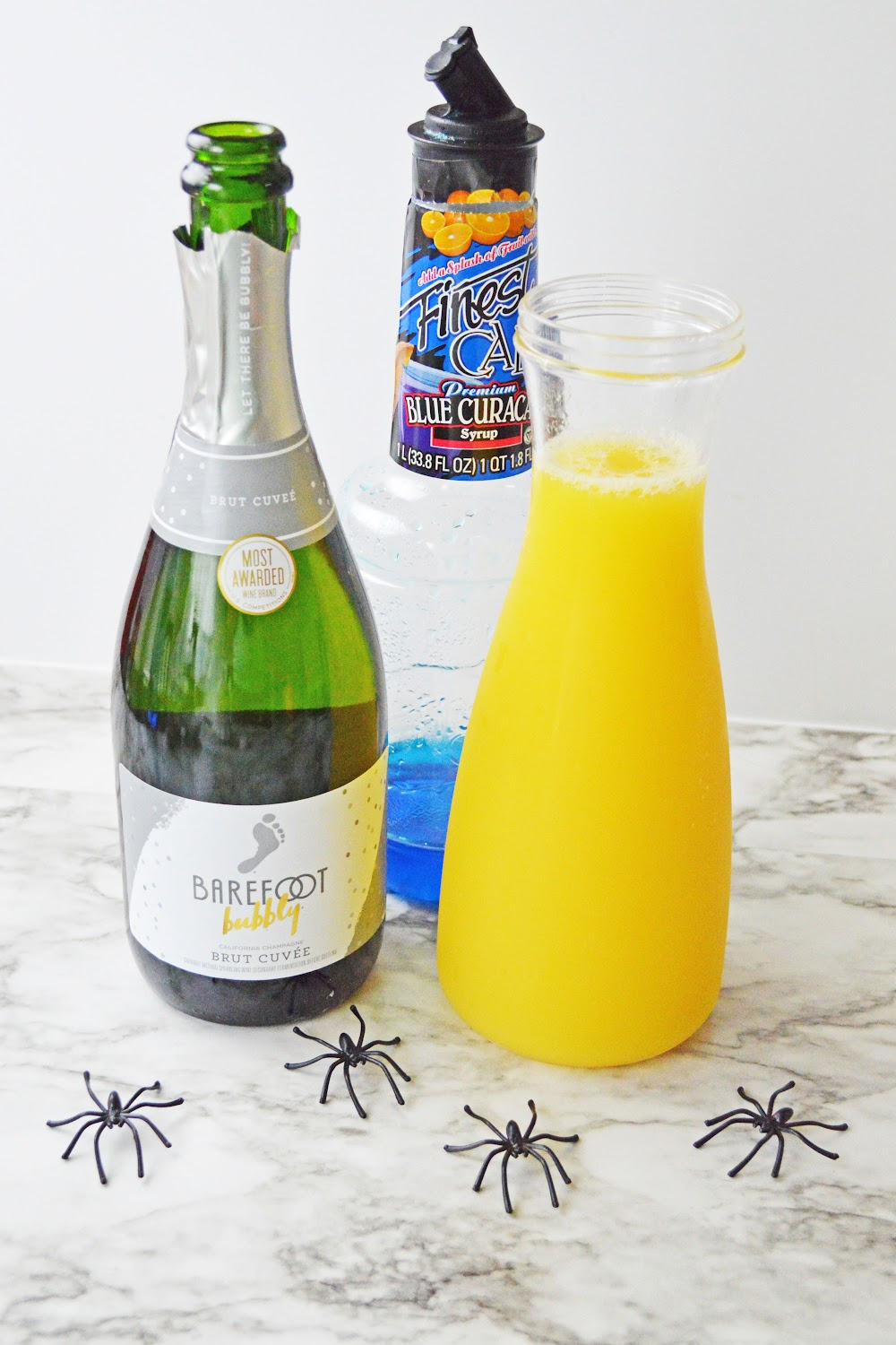Halloween Mimosas ingredients include champagne, orange juice and blue Curacao. These ingredients are displayed on a gray and white marble background with black plastic spiders.