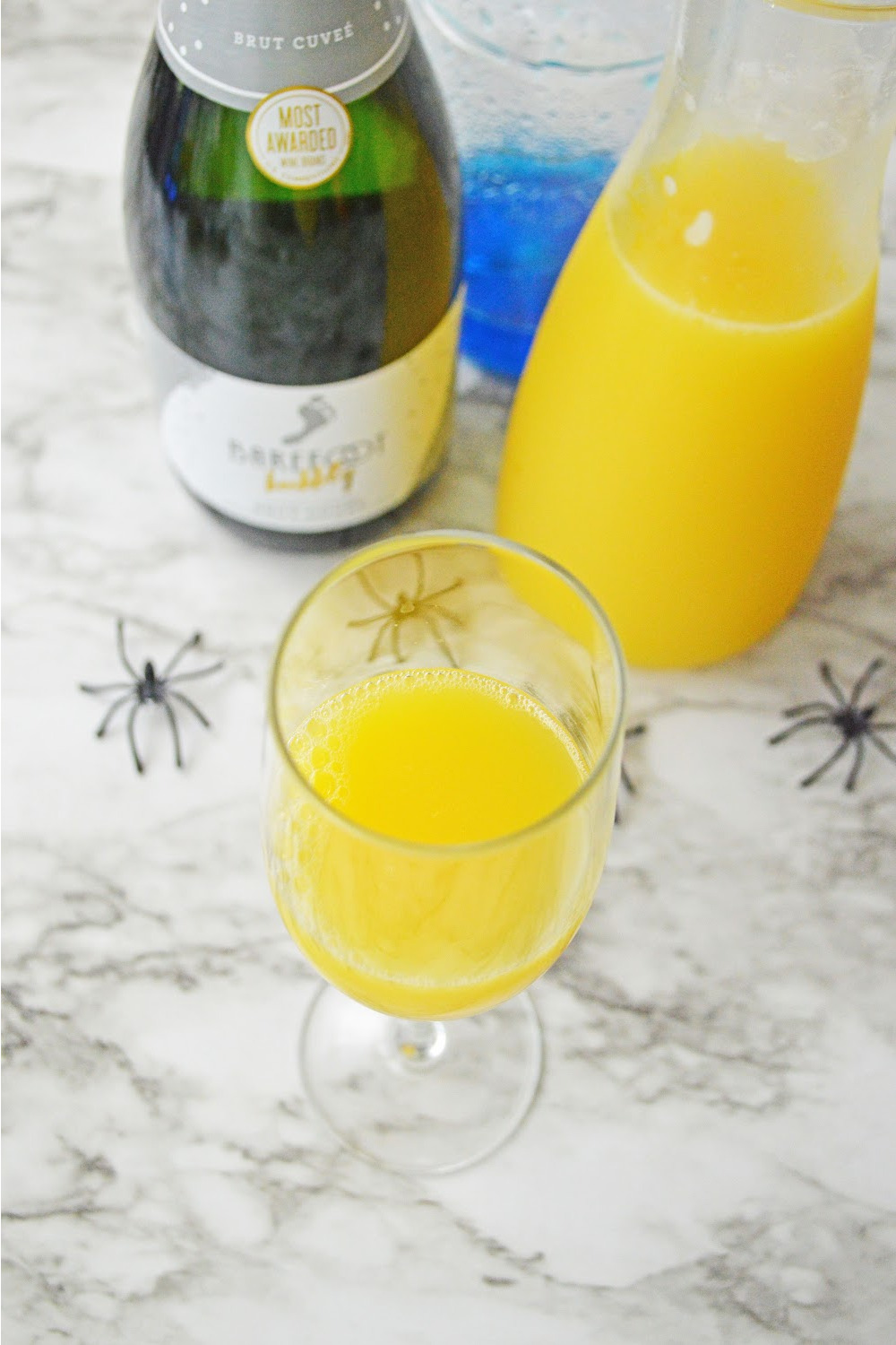Orange juice in a glass for theseHalloween Mimosas ingredients include champagne, orange juice and blue Curacao. These ingredients are displayed on a gray and white marble background with black plastic spiders.