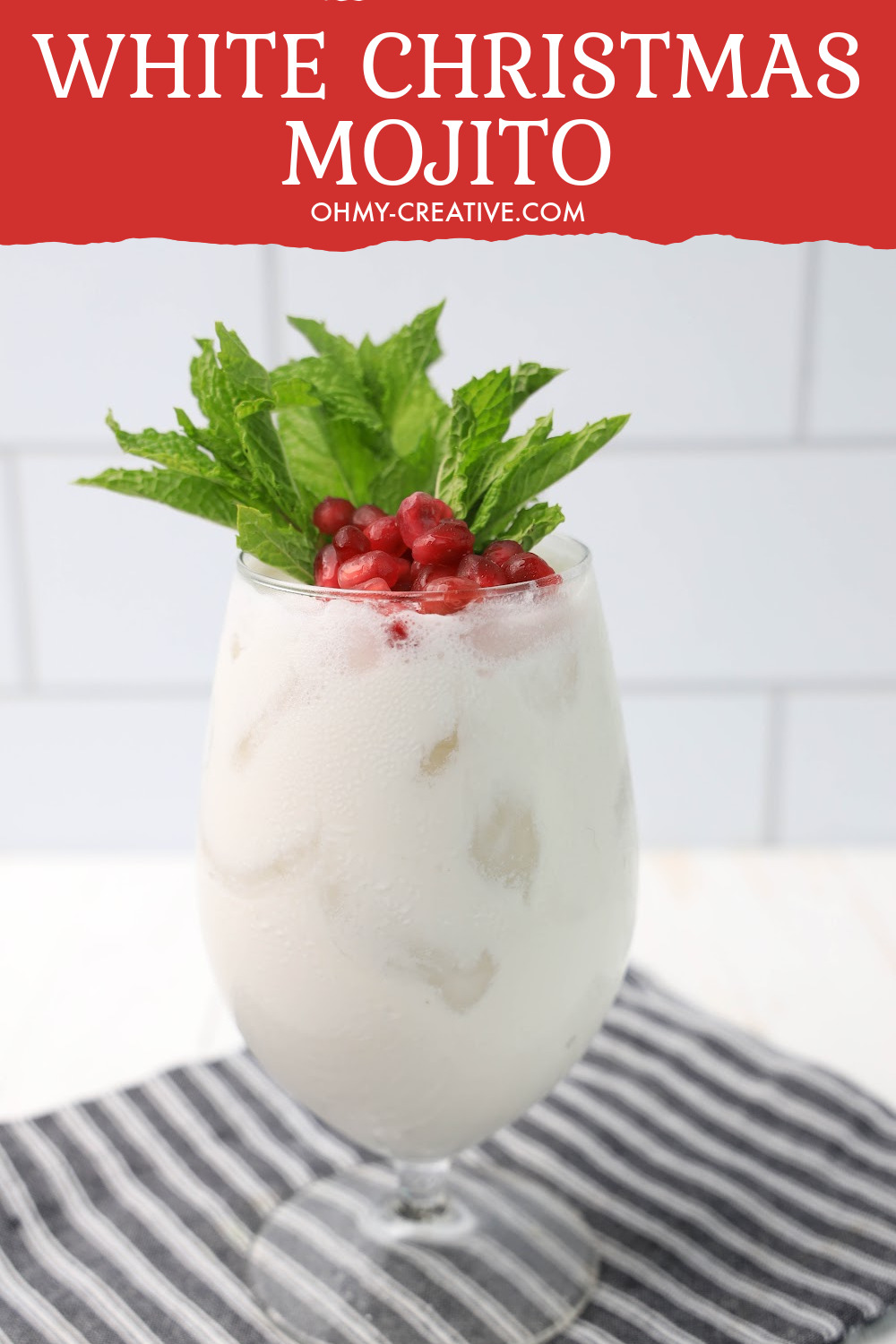 This pretty white Christmas mojito is topped with pomegranate arils and fresh mint. It sits on a black and white checked napkin on a white kitchen counter and backsplash.