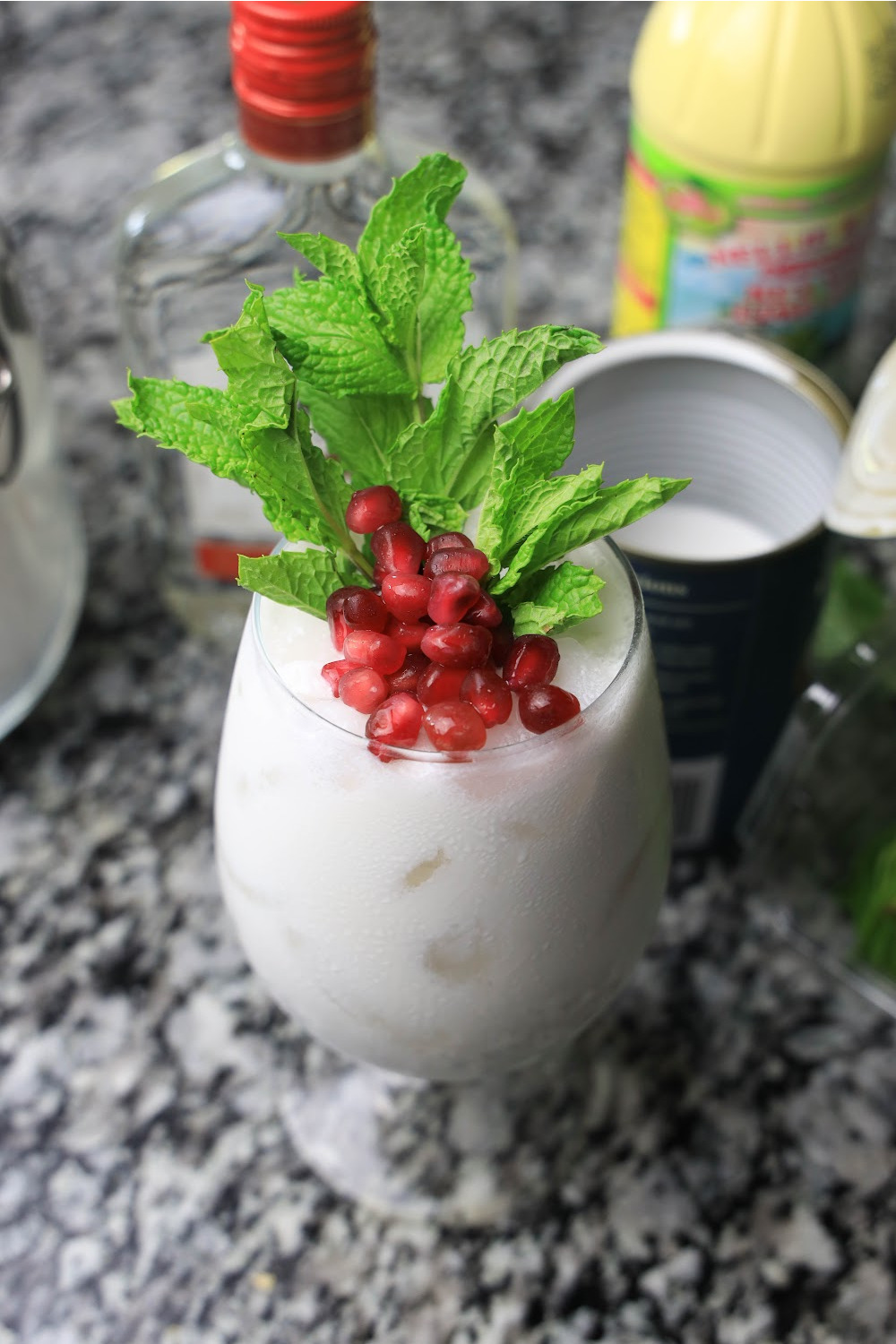 This pretty white Christmas mojito is topped with pomegranate arils and fresh mint. It sits on a black and white granite countertop with some of the mojito ingredients around it.