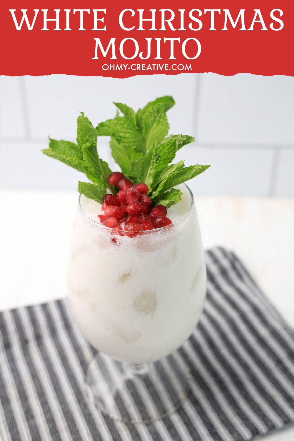 This pretty white Christmas mojito is topped with pomegranate arils and fresh mint. It sits on a black and white checked napkin on a white kitchen counter and backsplash.