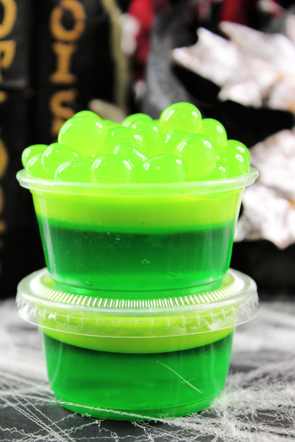 Bubbly Witches Brew Jello Shots for Halloween Parties. These green jello shots are sitting on a spiderweb background. The tops of the jello shots are topped with green apple popping bobas.