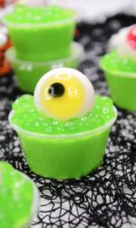 These Halloween Eyeball Jello Shots are topped with green boba and a gummy eyeball with yellow iris perfect for Halloween! Several of them are sitting on a black spiderweb background.