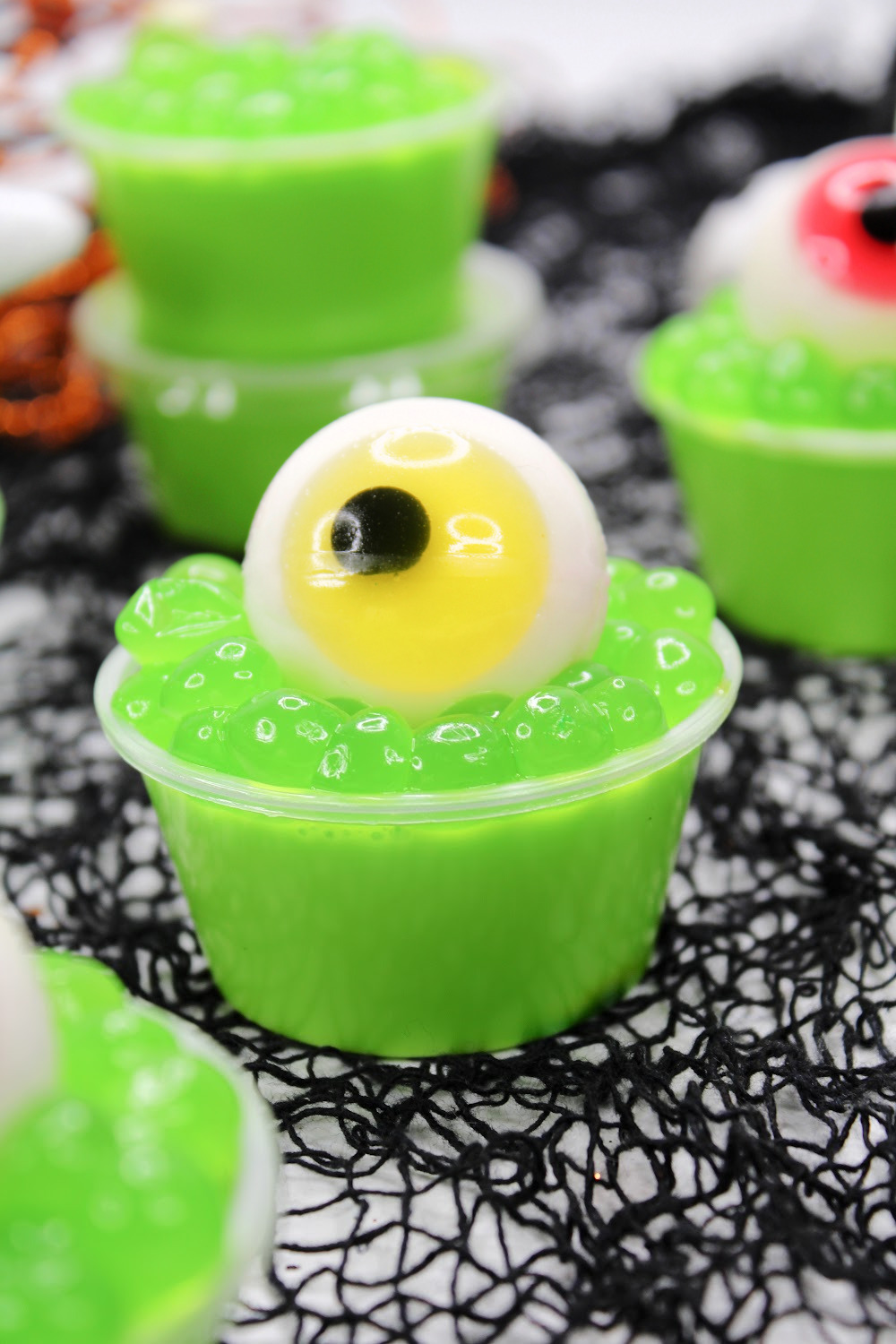 These Halloween Eyeball Jello Shots are topped with green boba and a gummy eyeball with yellow iris perfect for Halloween! Several of them are sitting on a black spiderweb background.