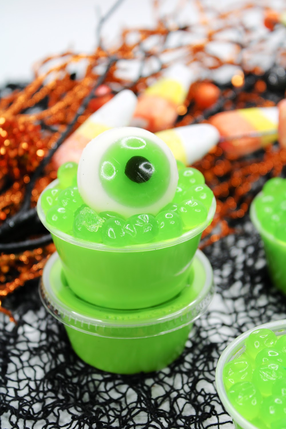 Stacked Halloween Eyeball Jello Shots are topped with green boba and a gummy eyeball with green iris perfect for Halloween! Several of them are sitting on a black spiderweb background.