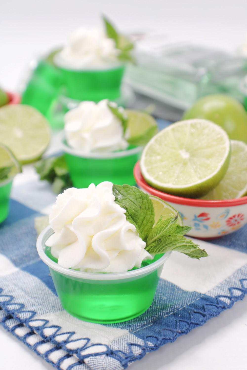 Mojito Jello Shots made with lime Jell-O and light rum. Garnished with whipped cream, a lime wedge and mint. These mojito jello shots are sitting on a blue and white checked cloth napkin with a bowl of sliced limes on the side.