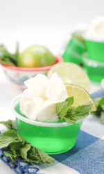 A close up of a Mojito Jello Shots made with lime Jell-O and light rum. Garnished with whipped cream, a lime wedge and mint. These mojito jello shots are sitting on a blue and white checked cloth napkin with a bowl of sliced limes on the side.