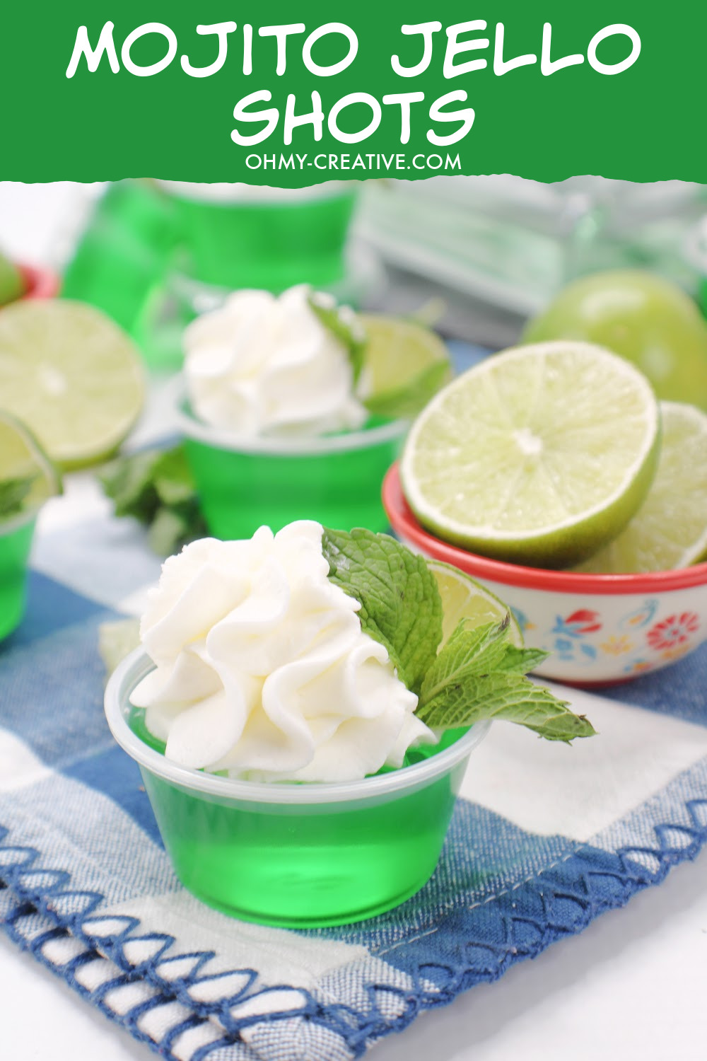 Mojito Jello Shots made with lime Jell-O and light rum. Garnished with whipped cream, a lime wedge and mint. These mojito jello shots are sitting on a blue and white checked cloth napkin with a bowl of sliced limes on the side.
