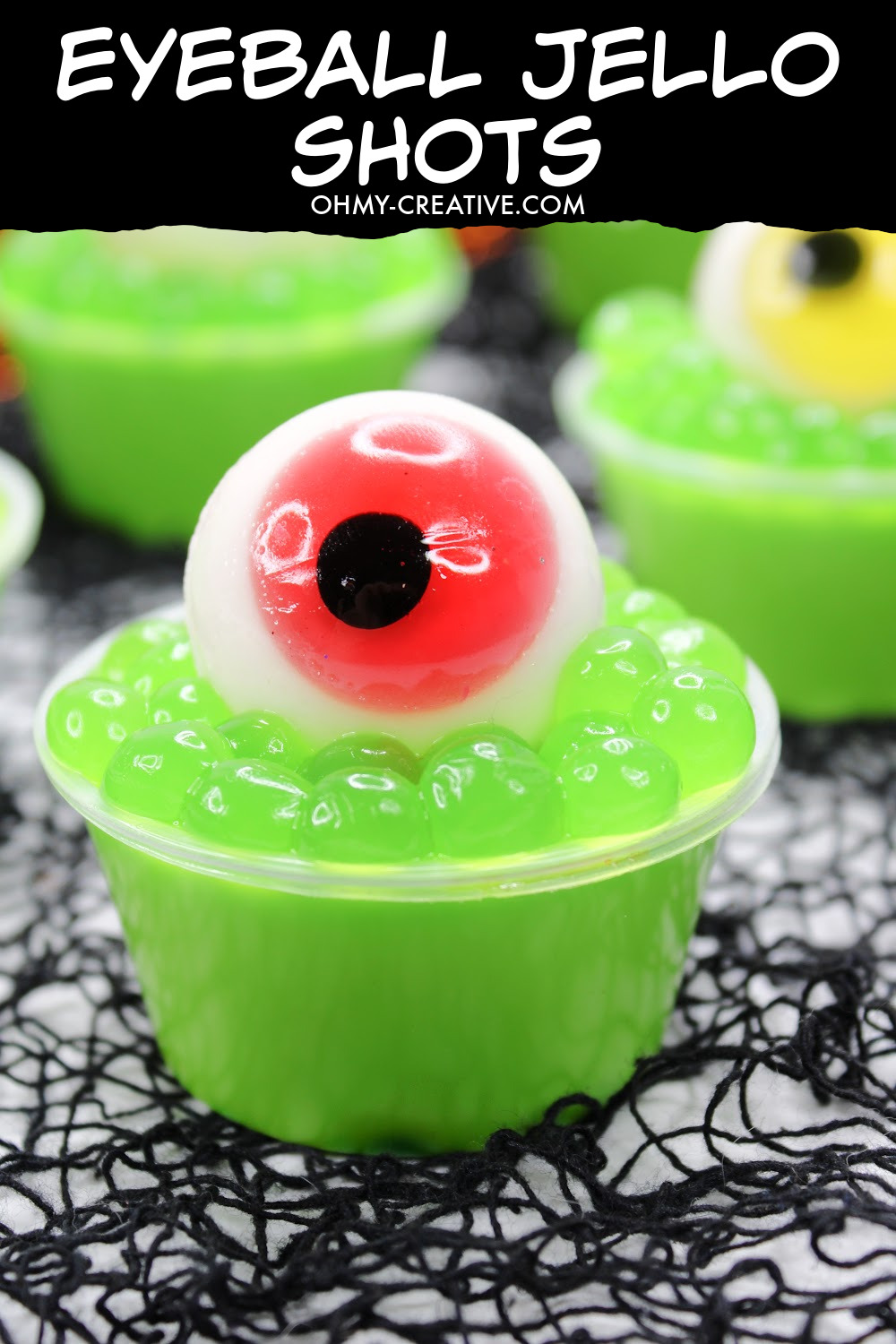 These Halloween Eyeball Jello Shots are topped with green boba and a gummy eyeball with red iris perfect for Halloween! Several of them are sitting on a black spiderweb background.