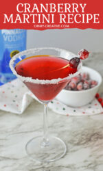 A cranberry martini with a sugar rim garnished with a skewer of sugar coated cranberries. Cranberry ingredients can be seen in the background.