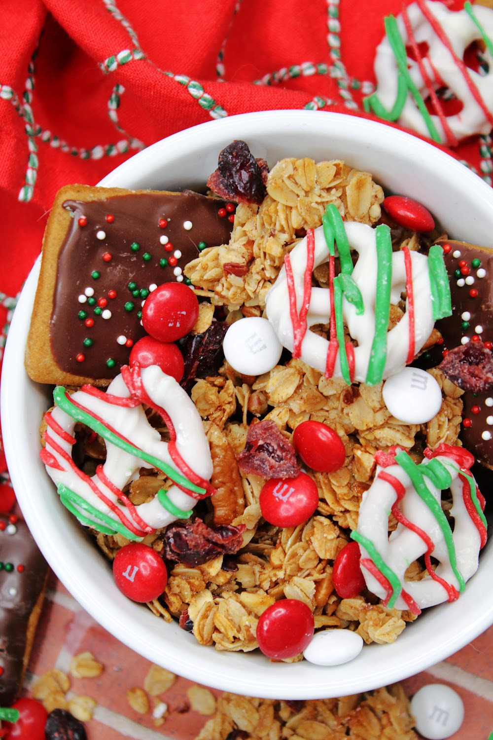 This reindeer chow includes gingerbread cookies, granola, M&Ms and white chocolate-covered pretzels. Served here in a white bowl with a festive red hand towel.