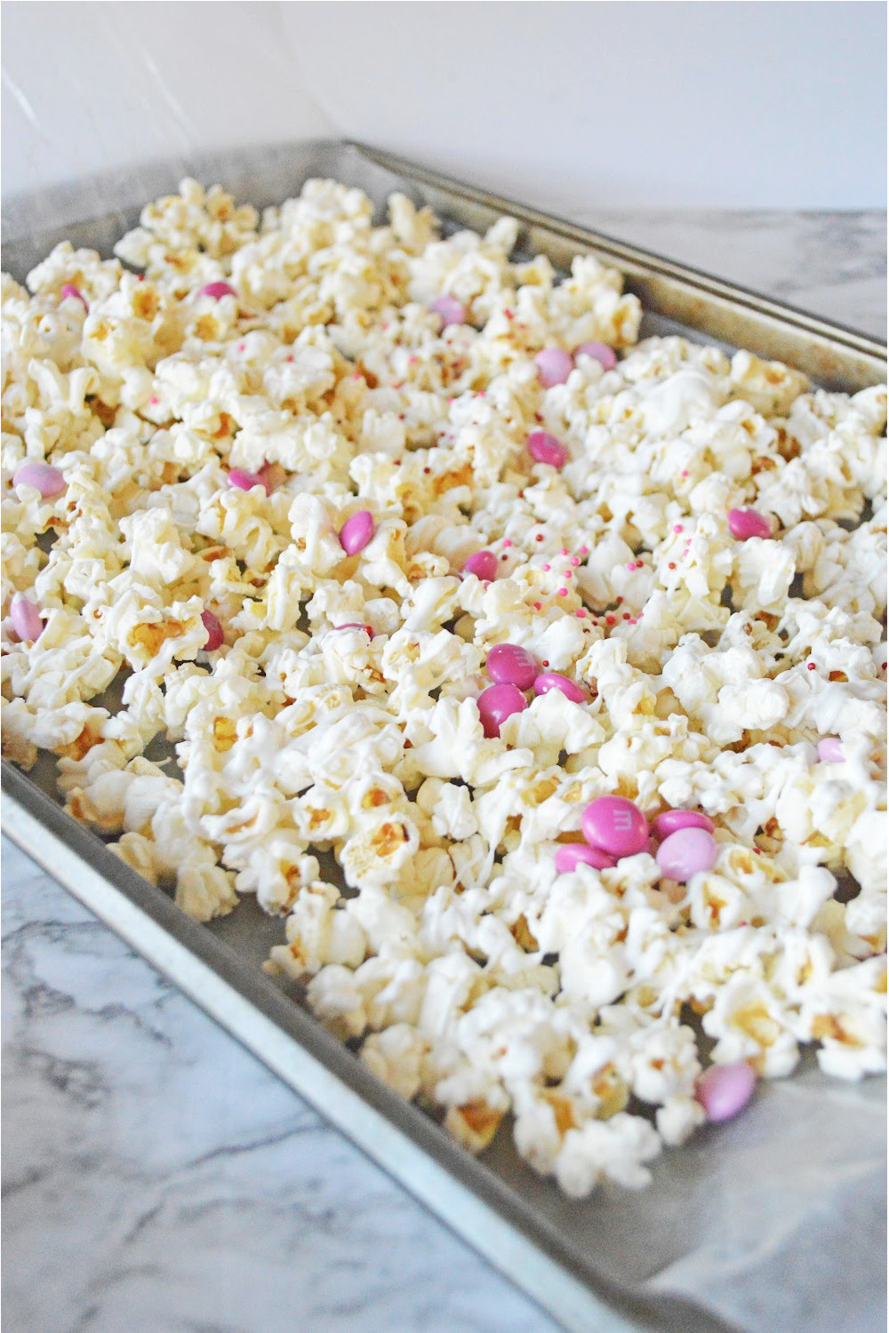 popcorn spread on a baking sheet with white candy melts drizzled on top plus pink M&Ms on top.