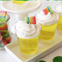 Pin image with Pot o' gold jello shots sitting on a white tray. Jar of rainbow candy sitting in the background.