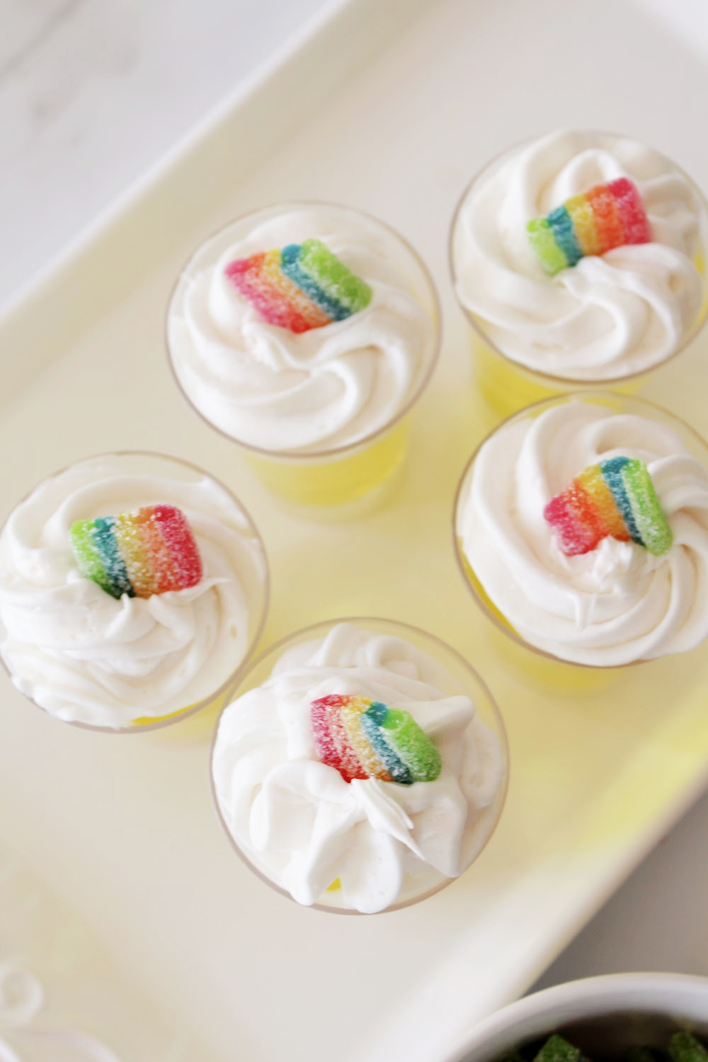 St. Patrick's Day jello shots topped with whipped cream and decorated with rainbow candy on top. Sitting on a white serving tray.