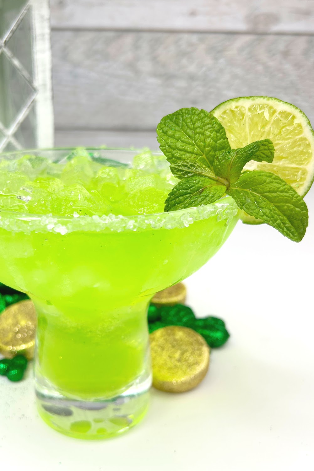 A salt rimmed green Irish margarita in a margarita glass garnished with a lime. In the background are shamrocks and gold pieces.