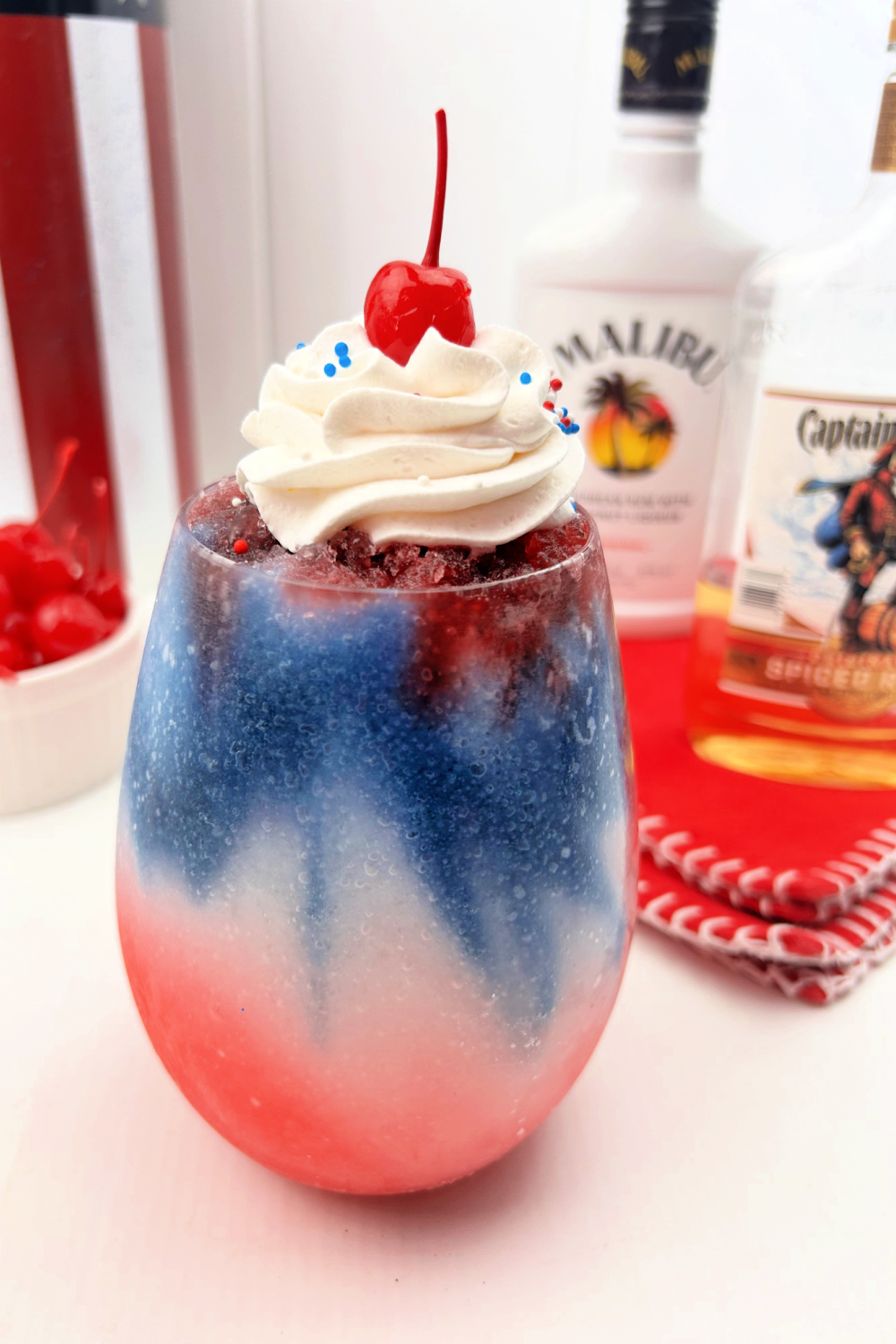 Featuring festive, patriotic colors, this Red White, And Blue Cocktail is like frozen slushy with red on the bottom and blue on the top layer with whipped cream and a cherry on top. Added are patriotic sprinkles. Bottle of the alcohol are in the background.