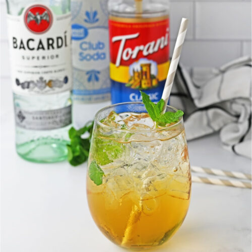 A caramel mojito cocktail garnished with mint and a straw. Alcohol bottles in the background.